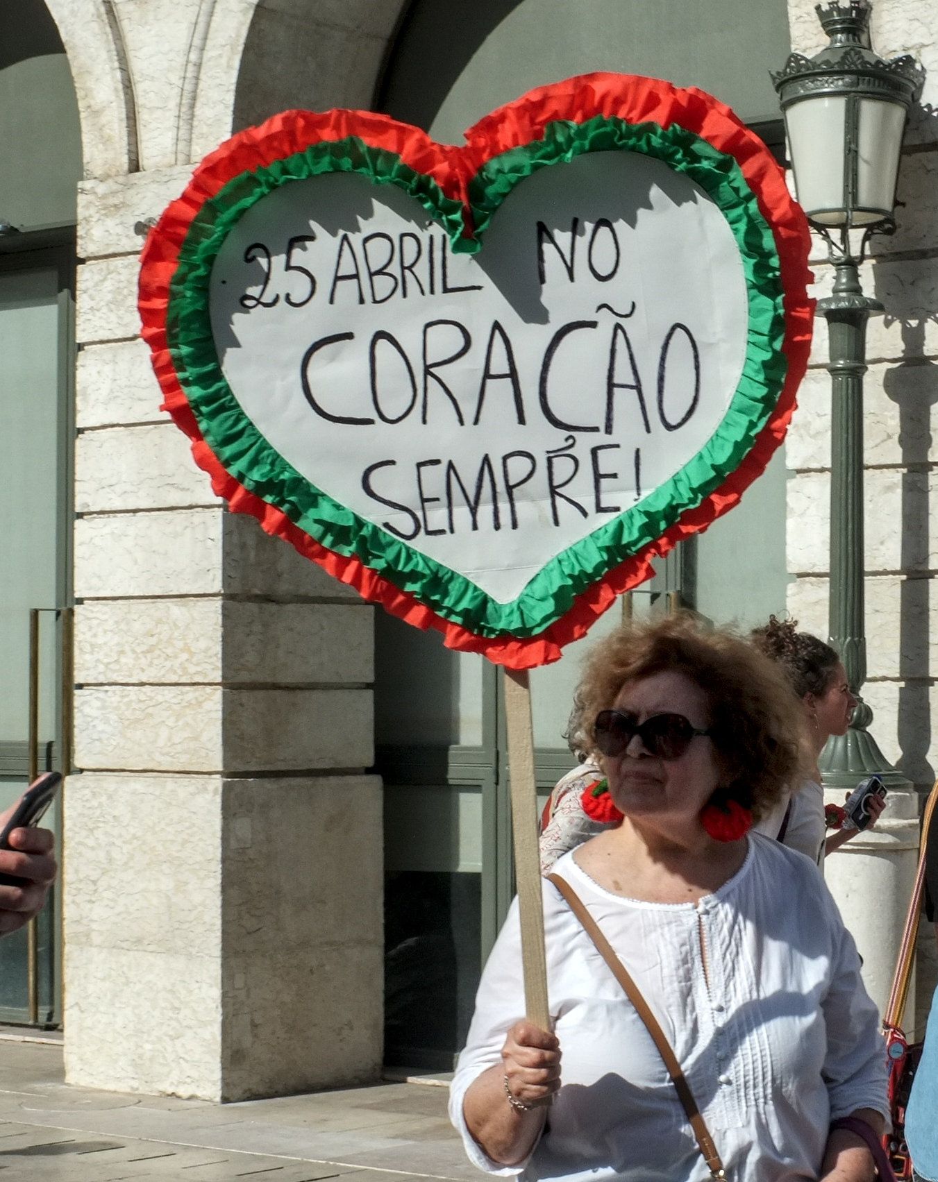 Uma senhora já com alguma idade, de óculos de sol e com dois brincos-cravos empunha um cartaz em forma de coração, com as cores verde e vermelho e a frase "25 Abril no Coração Sempre"