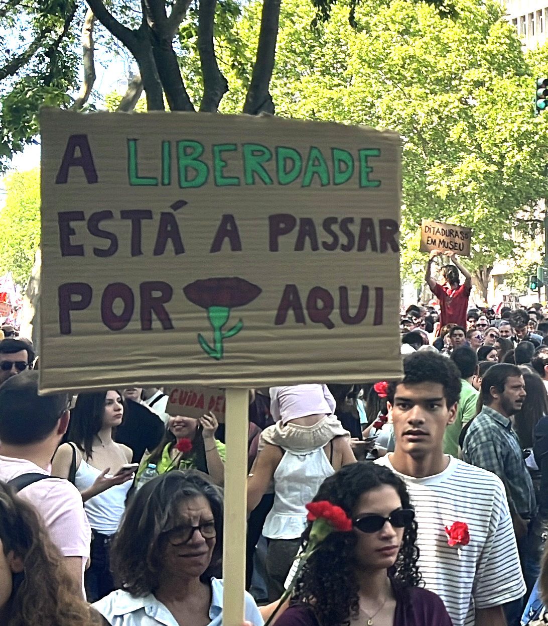 Uma pessoa no meio da multidão segura num cartaz que diz "A Liberdade está a passar por aqui", com um cravo desenhado"