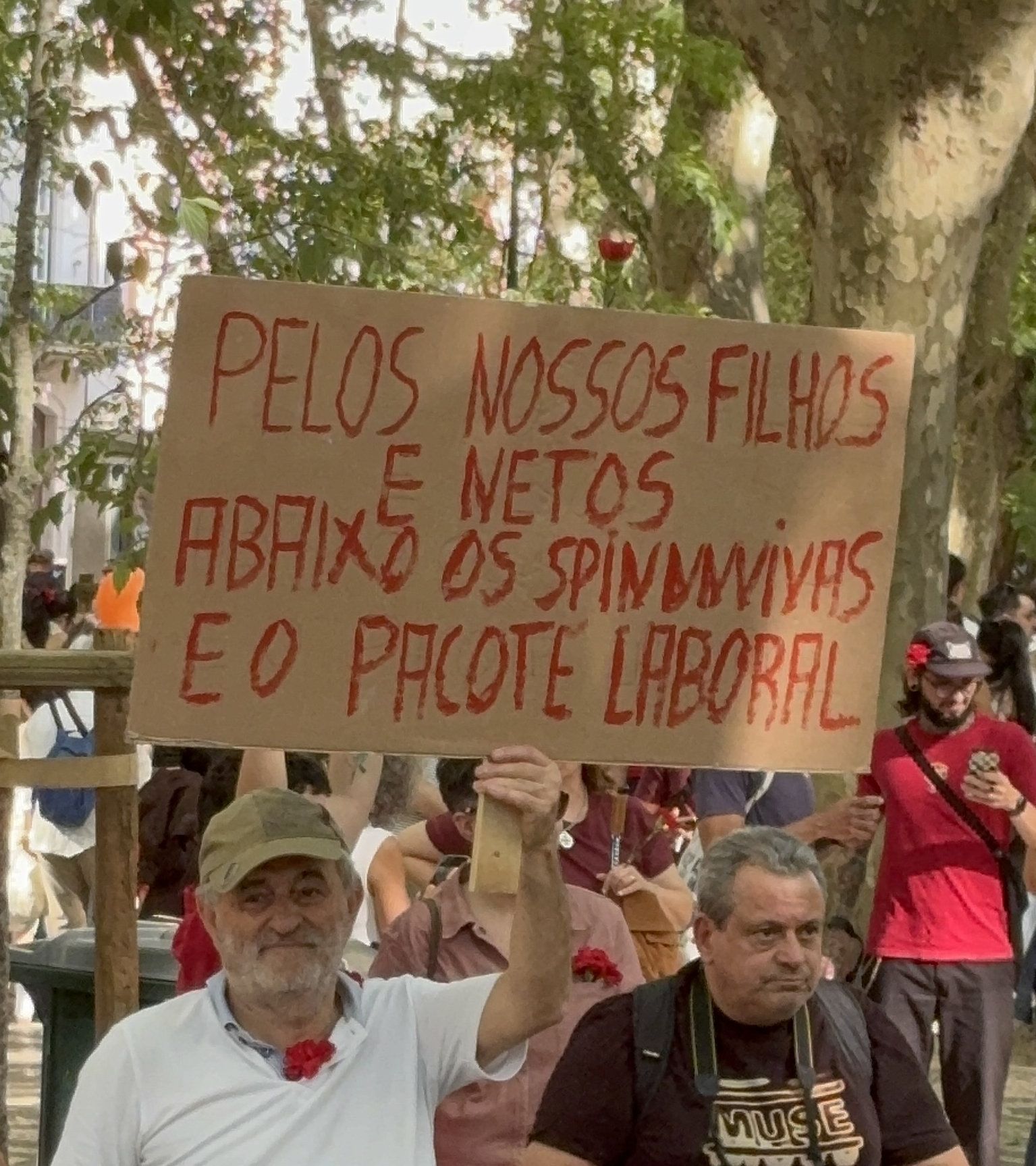 Um senhor vestido de t-shirt branca e com um cravo vermelho ao pescoço e um boné verde levanta um cartaz no qual se pode ler "Pelos nossos filhos e netos abaixo os Spinunvivas e o pacate laboral"."
