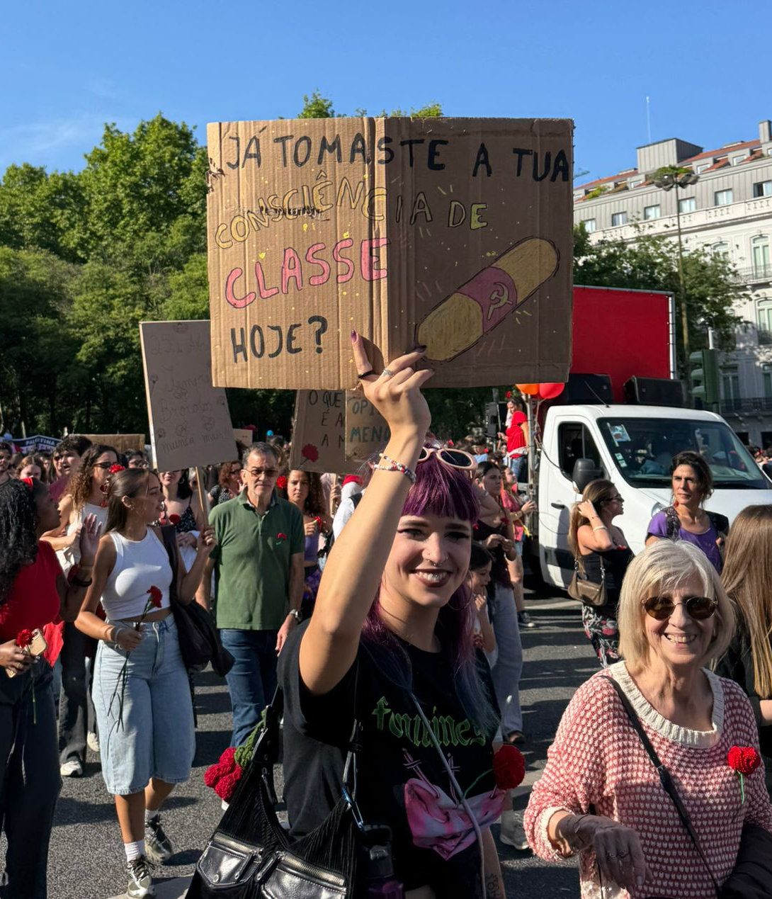 Uma jovem segura um cartaz no meio do desfile que contem as palavras "Já tomaste a tua consciência de classe hoje?" e inclui um comprimido comunista
