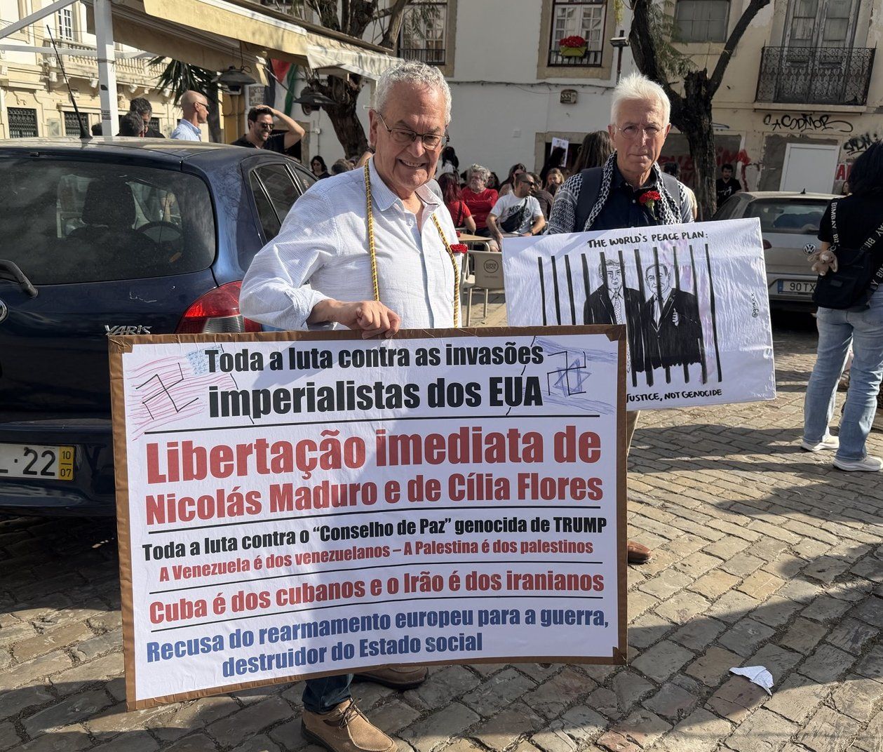 Dois senhores mais velhos seguram dois cartazes que denunciam o Imperialismo americano e Sionista em diferentes vertentes.
