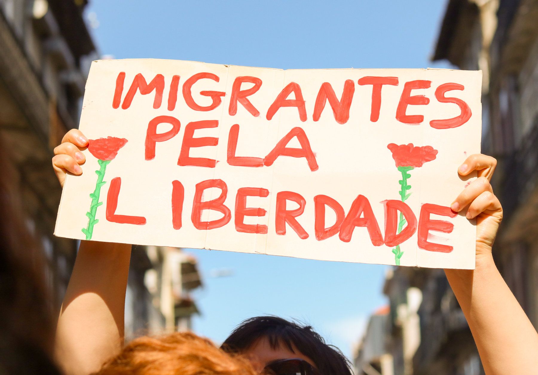 Uma pessoa segura um cartaz com letras vermelhas e dois cravos de cada lado. A frase diz "Imigrantes pela Liberdade".