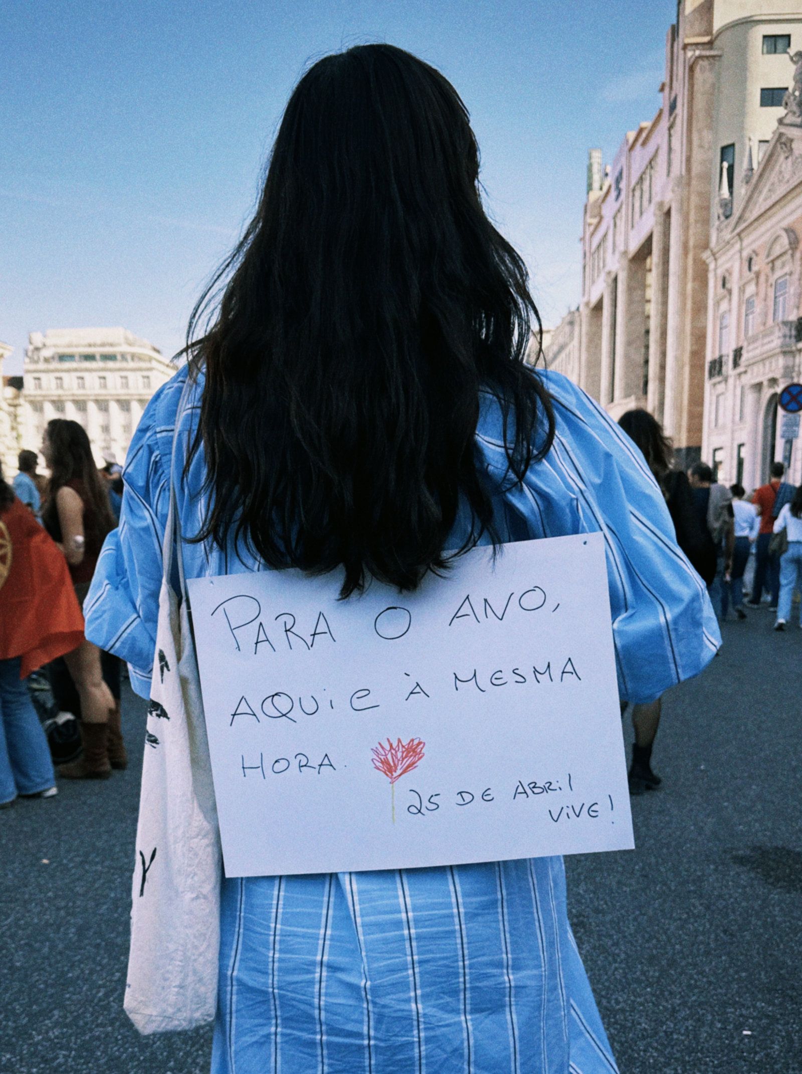 Uma rapariga de costas tem um sinal pendurado que diz "Para o Ano Aqui e à Mesma Hora"