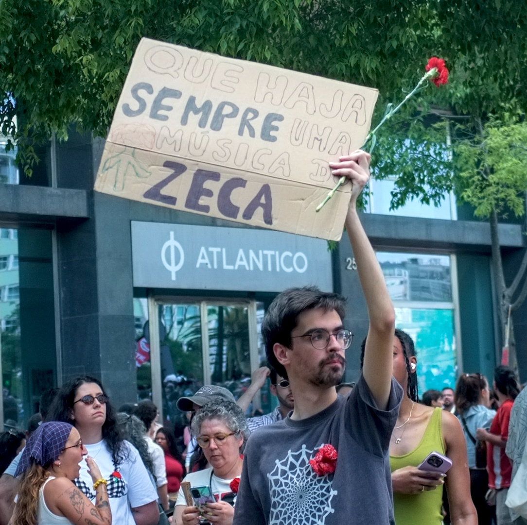 Um jovem segura um cartaz que diz "Que Haja Sempre uma Música do Zeca"