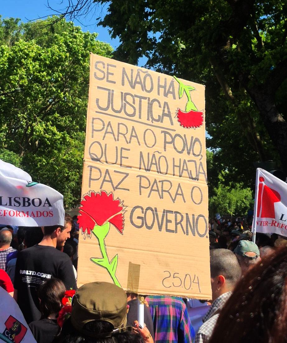 Um cartaz decorado com dois cravos diz "Que Não Haja Paz Para O Governo". Também tem a data, 25.04