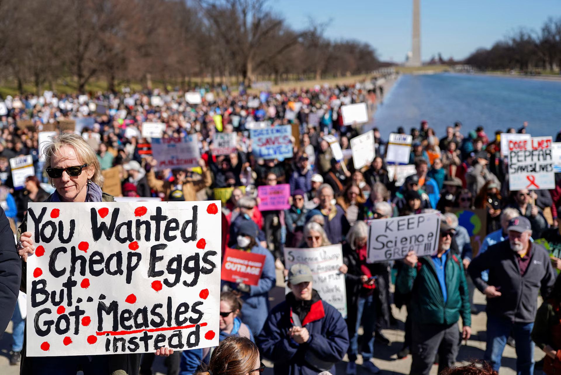 Uma senhora segura um cartaz que diz 'You Wanted Cheap Eggs, But...
      Got Measles instead.', com uma multidão de manifestante atrás, algures nos Estados Unidos.